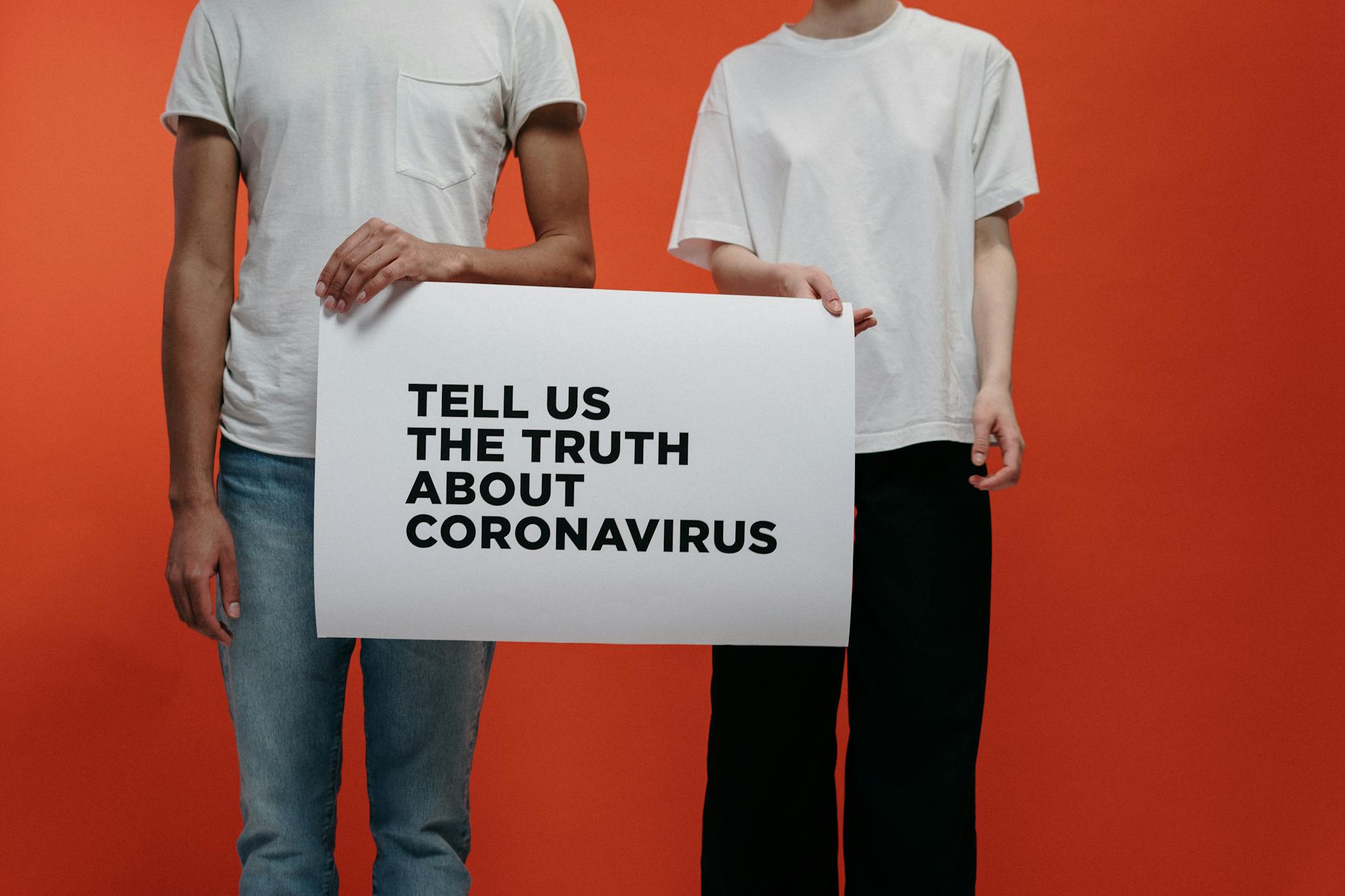 Two individuals holding sign demanding truth about coronavirus against orange background.
